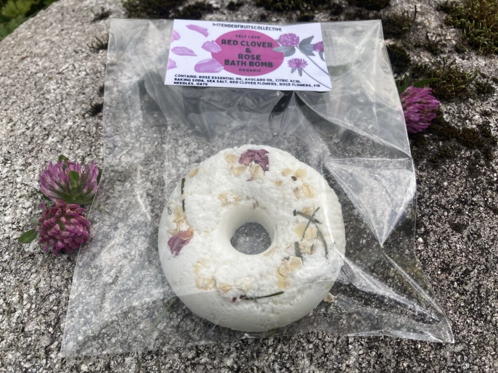a photo of a donut shaped bath bomb with fir needles, oats and rose petals on the top, sitting inside a clear bag with a tender fruits label on it, on top of a grey boulder, with a few red clover flowers next to the bag. The boulder is speckled and has dark black moss growing on them.