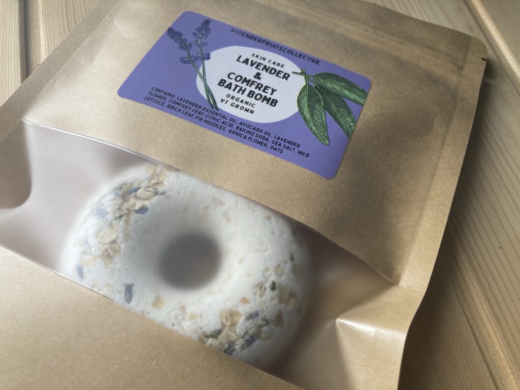 a photo of a donut shaped bath bomb with lavender flowers, oats and fir needles on the top, sitting inside a recycled craft-paper bag with a tender fruits label on it, is on top of a wooden surface. 