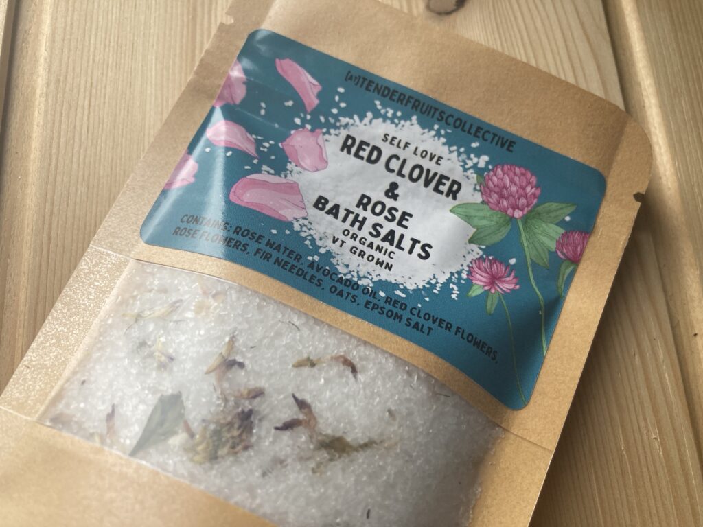 a photo of bath salts with fir needles, oats red clover flowers and rose petals in it, inside a sealed recycled craft-paper bag with a tender fruits label on it, is on top of a wooden surface.