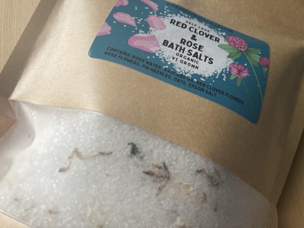 a photo of bath salts with fir needles, oats red clover flowers and rose petals on the top, inside a sealed recycled craft-paper bag with a tender fruits label on it, is on top of a wooden surface. 