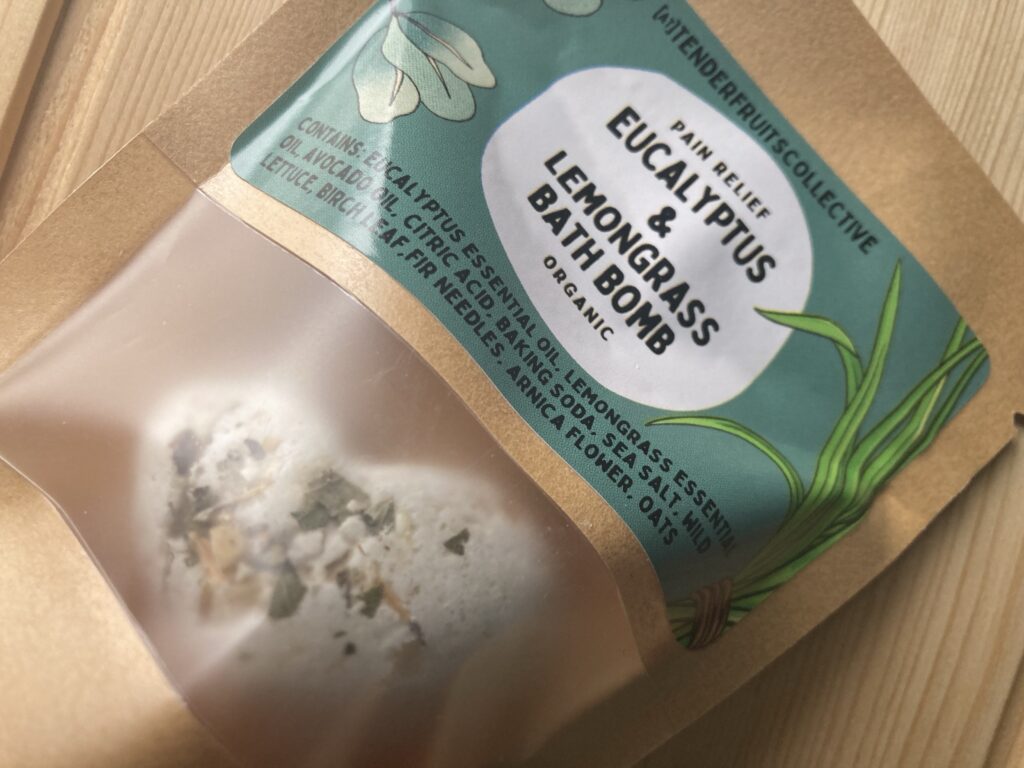 a photo of a heart shaped bath bomb with fir needles, oats and birch leaves on the top, sitting inside a recycled craft-paper bag with a tender fruits label on it, is on top of a wooden surface. 