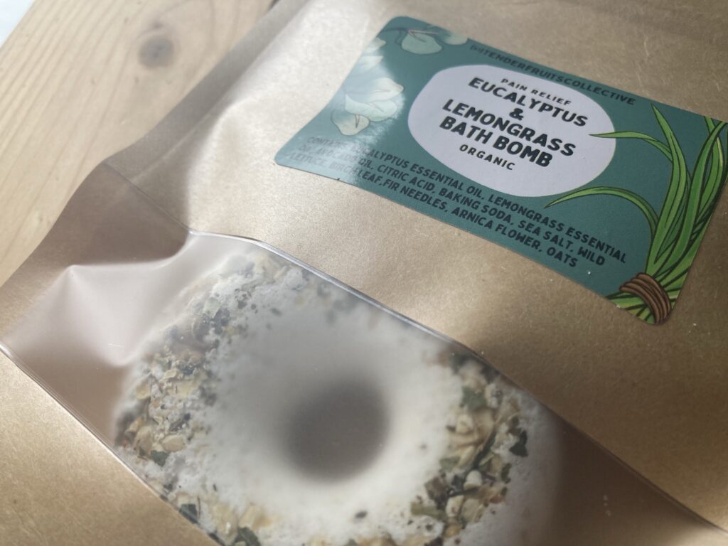 a photo of a donut shaped bath bomb with fir needles, oats and birch leaves on the top, sitting inside a recycled craft-paper bag with a tender fruits label on it, is on top of a wooden surface. 